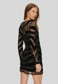Abito nero in pizzo con maniche lunghe, caratterizzato da un motivo a chevron di tessuti trasparenti e vellutati, e una chiusura con zip sul retro.