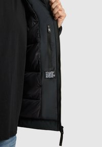 Ręka trzyma otwartą czarną pikowaną kurtkę, ukazującą wewnętrzną kieszeń na zamek oraz metkę z napisem "Khujo original changing threads."