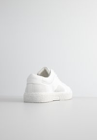 Zapatillas blancas con una suela de goma texturizada y ligeramente elevada. Cuerpo de cuero suave con detalles de malla y un sutil logo en el talón. Diseño minimalista.