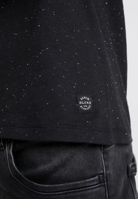 Camiseta negra con patrón salpicado blanco, de manga corta, y una etiqueta tejida que dice "Denim Blend with Attitude" en el dobladillo.
