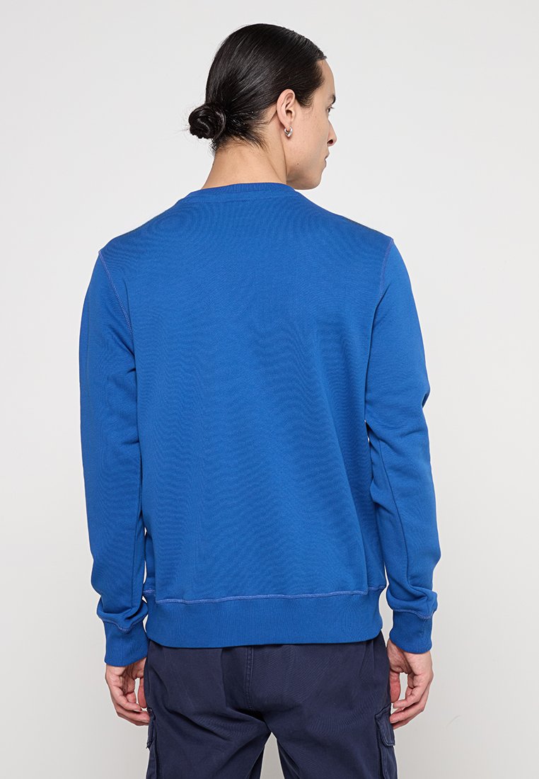 Uomo con capelli scuri raccolti in un chignon, che indossa una camicia a maniche lunghe blu e pantaloni scuri, in piedi con la schiena rivolta verso la fotocamera.