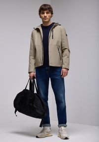 Veste imperméable beige avec capuche, deux poches avant, détails zippés noirs ; assortie à un jean bleu et un sac de sport noir.