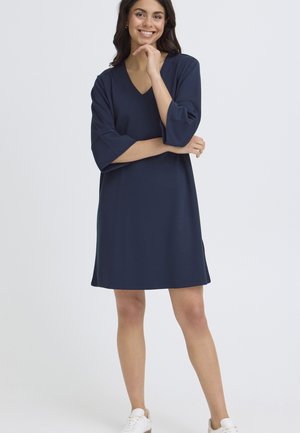 Jauna moteris šypsosi, vilki tamsiai mėlyną V formos kaklo suknelę su trimis ketvirčiais rankovių ir baltus sportinius batelius, stovi priešais paprastą foną.