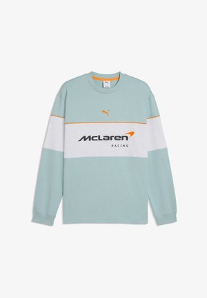 Chemise bleu clair à manches longues avec une rayure horizontale blanche, logo Puma orange au-dessus, et texte McLaren Racing en noir et orange sur la poitrine.