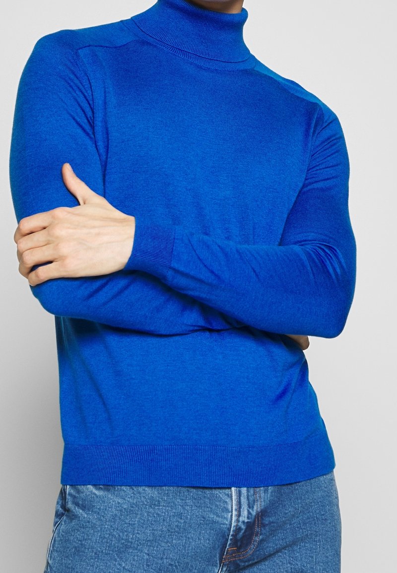 Blå polotröja i ett slätt stickat tyg, med ribbad krage och manschetter, figurnära design och diskreta sömdetaljer på axlarna.