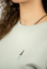 Hellgrünes Baumwoll-T-Shirt mit einem kleinen schwarzen Baumdesign auf der linken Seite. Der Stoff hat eine glatte Textur und einen Rundhalsausschnitt.