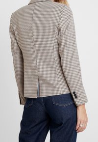 Kratkowana marynarka w beżowo-czarnym materiale, o dopasowanym kroju, z jednym rozcięciem z tyłu, mankietami zapinanymi na trzy guziki i subtelnym paskiem na dole.