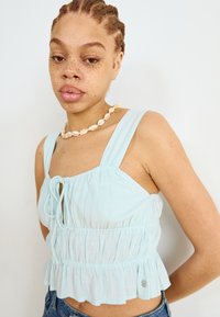 Lyseblå kort topp med justerbar knytning, samlet tekstur og volangkant. Bært med et skjell-halskjede og denimshorts.