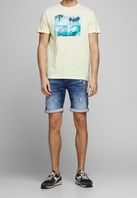 Hombre con una camiseta amarilla pálida con un gráfico de playa azul, pantalones cortos de mezclilla desgastados y enrollados, y zapatillas negras y blancas, de pie sobre un fondo gris.