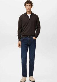 Junger Mann steht mit einer Hand in der Tasche, trägt einen dunkelbraunen Pullover mit Reißverschluss am Hals, blaue Jeans und beige Turnschuhe vor einfachem Hintergrund.