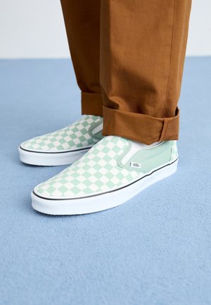Pieds portant des baskets Vans slip-on à damier vert et blanc, associées à un pantalon marron retroussé sur un sol bleu clair.