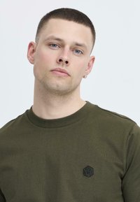 Pulover verde măsliniu, cu guler rotund, cu mansete ribbate, având o mică etichetă cu logo negru pe piept, cu o textură netedă și moale.