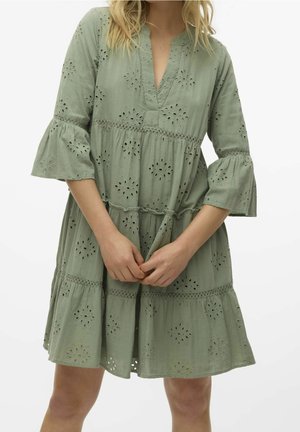 Woman wearing a light green, knee-length dress with floral eyelet patterns and flared three-quarter sleeves, standing with hands clasped.