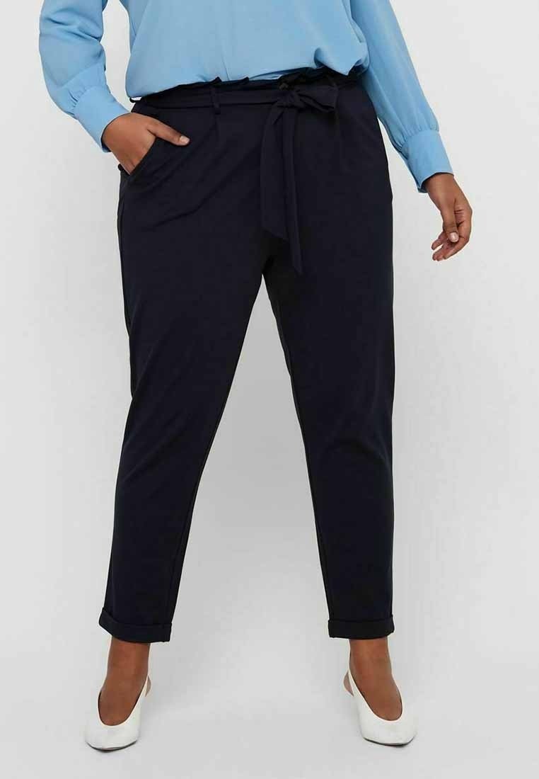 Person wearing black tapered pants with a tied waist belt, light blue blouse, and white closed-toe heels standing with one hand in pocket.