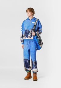 Chándal azul con estampado, que incluye un pulóver con cierre de cremallera hasta el cuarto y pantalones a juego. Diseñado con imágenes escénicas, complementado con un bolso de hombro azul y botas marrones.