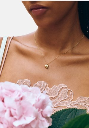 Gouden hartvormige hangerketting aan een dunne ketting, gedragen met een lichtroze kanten top. Omgeven door roze hortensia bloemen en groene bladeren.