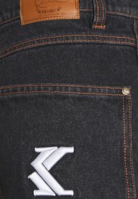 Mörka jeans i denim med orange sömmar, vitt broderat märke på bakfickan och en brun läderetikett vid midjan.