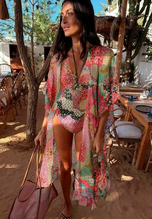 Femme en maillot de bain coloré à motifs et paréo assorti tenant un sac, debout dans un espace repas extérieur ombragé avec des meubles en bois.