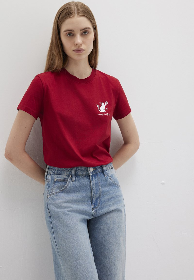 Red cotton T-shirt with short sleeves, featuring a white graphic design on the left chest. Paired with light blue high-waisted jeans.