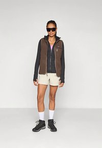 Γυναίκα φορώντας καφέ φλις γιλέκο, μαύρο αδιάβροχο μπουφάν με φερμουάρ, μπεζ σορτς, άσπρες κάλτσες, μαύρες ορειβατικές μπότες και μεγάλες μαύρες ηλιοφάνες, στέκεται σε απλό φόντο.