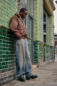 Brown padded jacket with a shearling collar, loose faded blue jeans, and black chunky shoes, leaning against a green tiled wall.