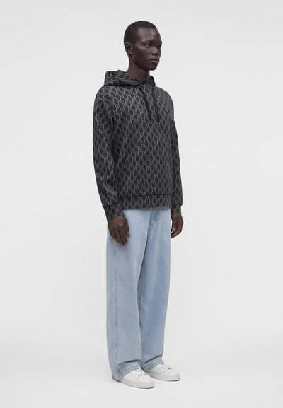 Homme portant un sweat à capuche noir à motifs, un jean bleu clair à jambes larges et des baskets blanches, debout devant un fond clair uni.