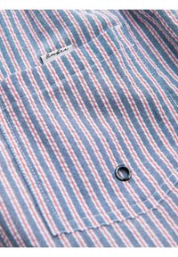 Tissu rayé bleu, blanc et rouge avec une poche à bouton et une petite étiquette cousue près du haut de la poche.