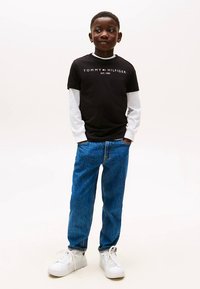 Zwart T-shirt met witte lange mouwen, blauwe spijkerbroek en witte sneakers. Het T-shirt heeft het logo "TOMMY HILFIGER" en het oprichtingsjaar.