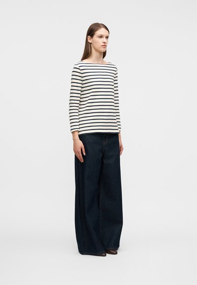 Νεαρή γυναίκα που στέκεται, φορώντας λευκό μπλουζάκι με μαύρες οριζόντιες ρίγες και σκούρο, φαρδύ παντελόνι τζιν, πάνω σε απλό φόντο.