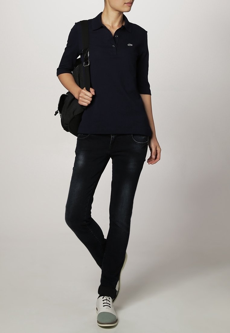 Personne portant un polo noir à manches trois-quarts, un jean skinny foncé, des baskets blanches et grises, portant un sac à dos noir sur une épaule.