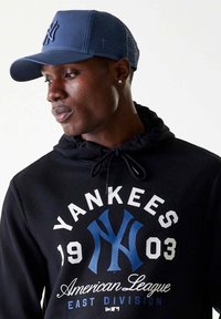 Felpa nera con cappuccio, caratterizzata da un logo e testo dei Yankees in bianco e blu. Cappello blu con retro in rete, che mostra il logo dei Yankees in blu scuro.
