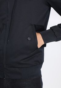 Black bomber jacket with a zip closure, ribbed cuffs, and a side pocket featuring a button detail. Smooth fabric texture.