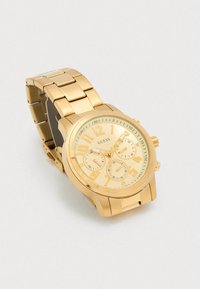 Relógio de pulso em metal dourado com bracelete ligada, mostrador circular, três subdials e marcadores de horas com números em relevo, exibindo o nome da marca Guess.
