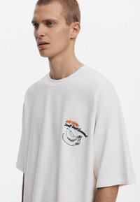 Tricou alb oversized cu mâneci scurte, având un grafic de banană negru și portocaliu cu text în zona stângă a pieptului. Textură de țesătură moale.
