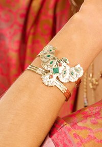 Gold- und Silberschnalle mit einem aufwändigen Vogeldesign, das grüne und rosa Steine enthält, vor einem bunten, gemusterten Stoffhintergrund.