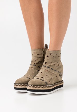 Bottines en suede khaki avec des motifs découpés, des accents noirs, un contour en jute tissé et une semelle compensée. Elles sont dotées d'une fermeture éclair sur le côté.