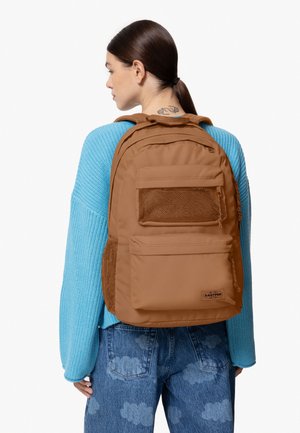 Zaino in tessuto marrone con accenti in rete e più tasche frontali, dotato di un patch con logo. Indossato sopra un maglione azzurro chiaro con maniche in tessuto testurizzato.