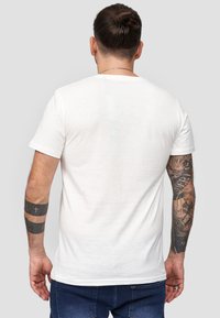 Weißes Baumwoll-T-Shirt mit kurzen Ärmeln, schlanker Schnitt, schlichtes Design. Auffällige Tattoos auf den Armen, mit leichtem Muster.