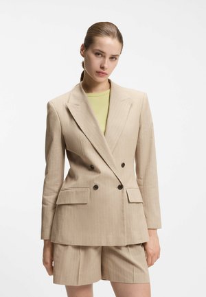 Kobieta z gładko zaczesanymi włosami, ubrana w beżową, dwurzędową marynarkę w prążki, na jasnokołnierzykowej bluzce w jasnej zieleni i dopasowanych szortach, na tle białego tła.