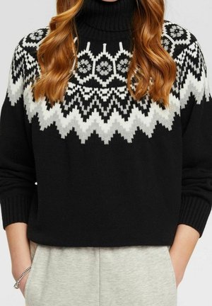 Femme aux cheveux roux ondulés portant un pull à col roulé noir avec des motifs géométriques blancs et gris sur la poitrine et les épaules.