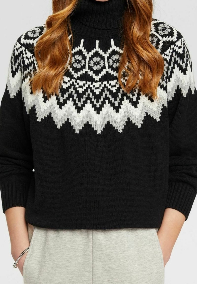 Femme aux cheveux roux ondulés portant un pull à col roulé noir avec des motifs géométriques blancs et gris sur la poitrine et les épaules.