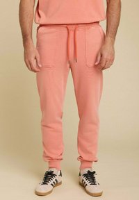 Pantalons de survêtement en coton rose avec taille élastique et cordon de serrage. Comprend deux poches avant et des poignets côtelés. Portés avec des baskets noires et blanches.