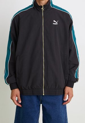 Homme portant une veste zippée noire Puma avec des rayures bleu sarcelle et blanches sur les manches et un jean bleu, debout les bras détendus.