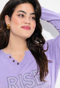 Camicia a maniche lunghe lilla con scollo a V, caratterizzata da tre bottoni decorativi e la scritta "RISE" stampata in strass. Gli orecchini a cerchio dorati completano il look.