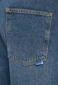 Denimstoff med en dyp blåfarge, med en rektangulær baklomme akkompagnert av oransje søm og en liten blå etikett ved sømmen.