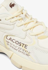 Baskets Lacoste en cuir blanc cassé avec des accents beiges. Dotées de coutures ton sur ton, de lacets plats et de coordonnées géographiques imprimées sur le côté.
