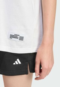 Bijela pamučna majica s etiketom koja prikazuje crni tekst i crne šortseve s bijelim Adidas logotipom. Majica ima opušten kroj i bočne proreze.