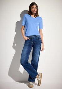 Svetle modré tričko s krátkým rukávem a výstřihem do V v kombinaci s tmavě modrými kalhotami s širokými nohavicemi. Model má na sobě béžové tenisky s krémovými akcenty. Jednoduché pozadí.