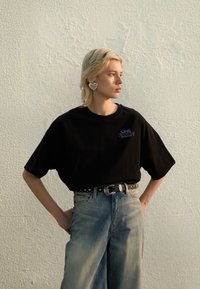 Sort oversized t-skjorte med lilla bokstaver, kombinert med lyse, brede jeans og et svart belte med nagler, med et sølvfarget øredobb formet som et hjerte.