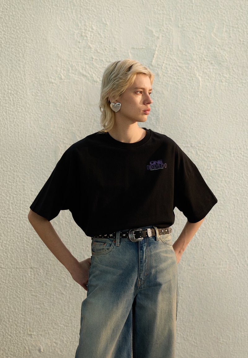Sort oversized t-skjorte med lilla bokstaver, kombinert med lyse, brede jeans og et svart belte med nagler, med et sølvfarget øredobb formet som et hjerte.